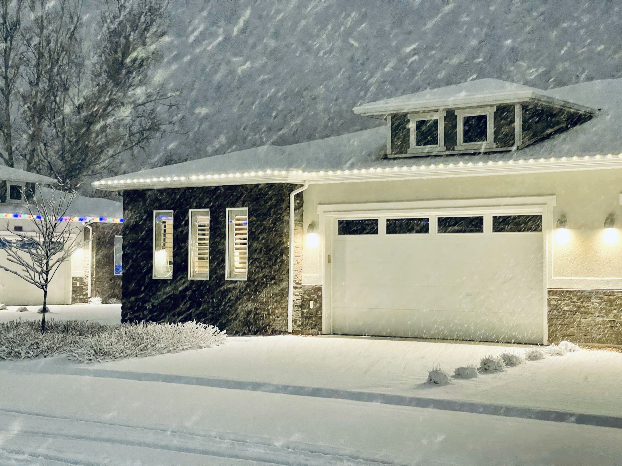 A home with holiday lights in a neighborhood during an evening snowstorm.